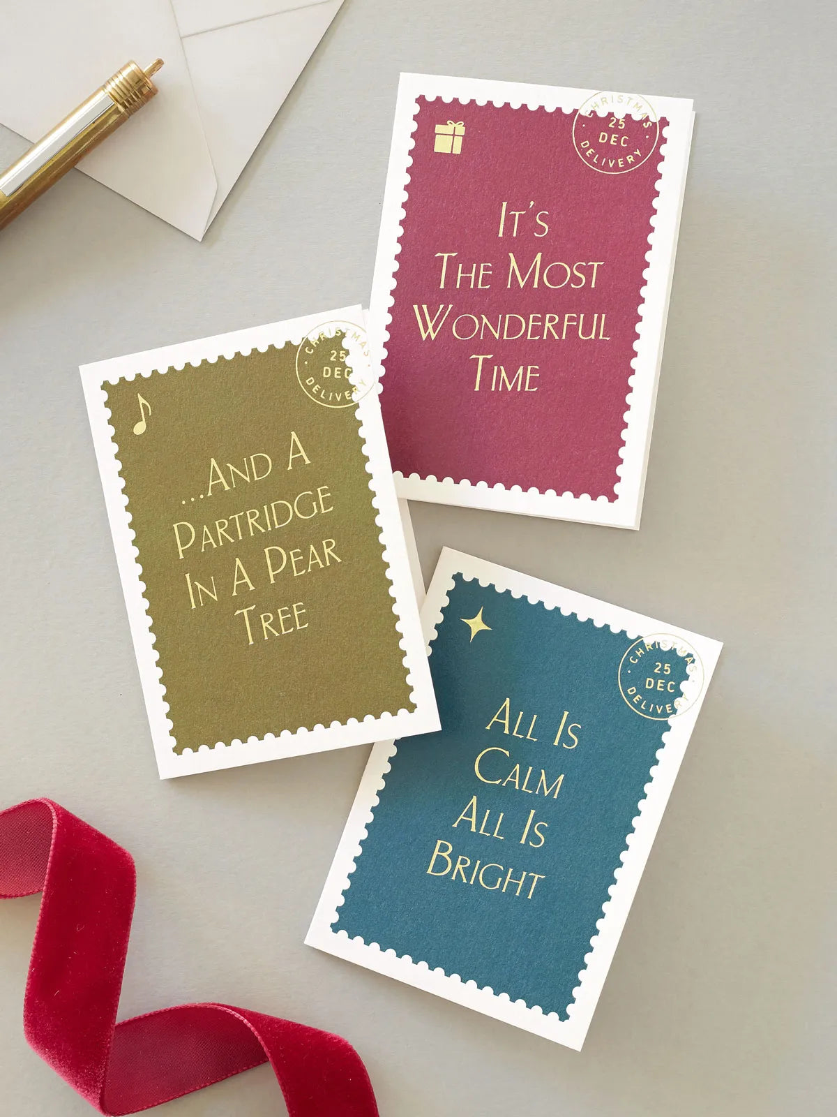 Three mini postage-stamp-style greeting cards with gold hot foil text are arranged on a light grey surface with a red ribbon, grey envelope, and gold pen as props.