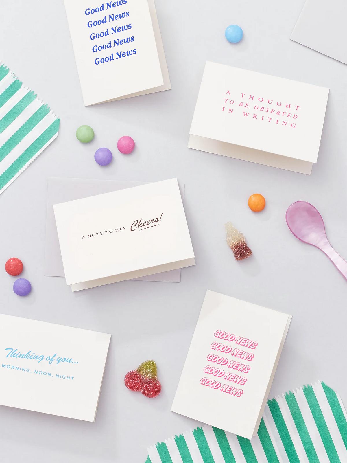 A styled flat-lay of several white greeting cards with pink, blue, and gold hot foiled text, arranged on a light gray surface with colorful sweets, a pink spoon and striped paper bag.