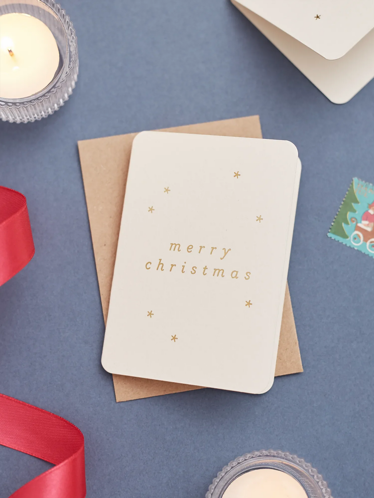 A minimalist mini Christmas greeting card with a warm off white card stock with gold foil text reading 'merry Christmas' and small gold stars, resting on a brown kraft envelope. The card is styled on a blue surface with postage stamps and ribbon around.