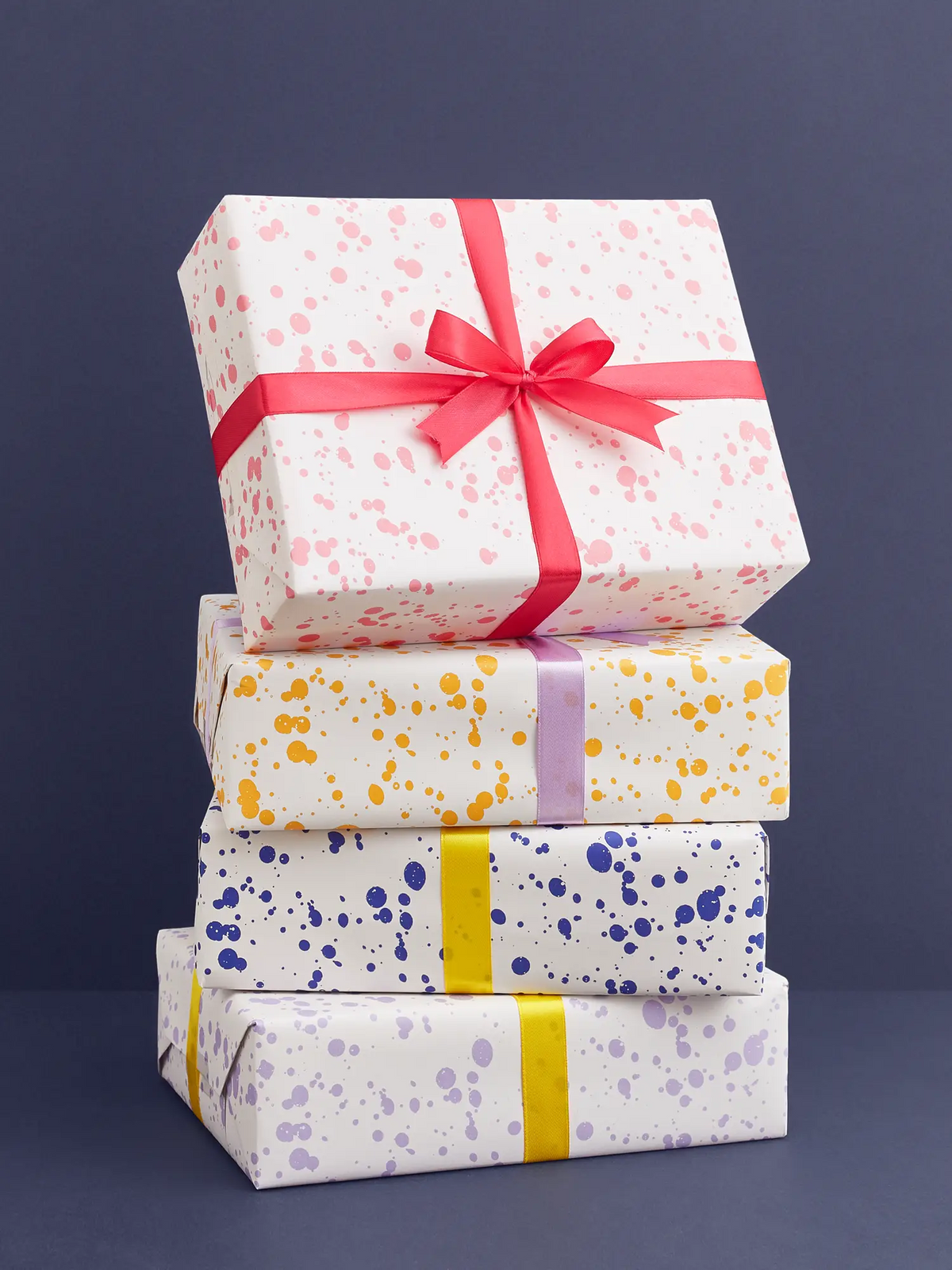 A stack of four wrapped gift boxes on a dark blue background. The boxes are white with colorful paint splatter patterns in pink, orange, blue, and lavender, each tied with a contrasting ribbon in red, yellow, lavender, and light purple.