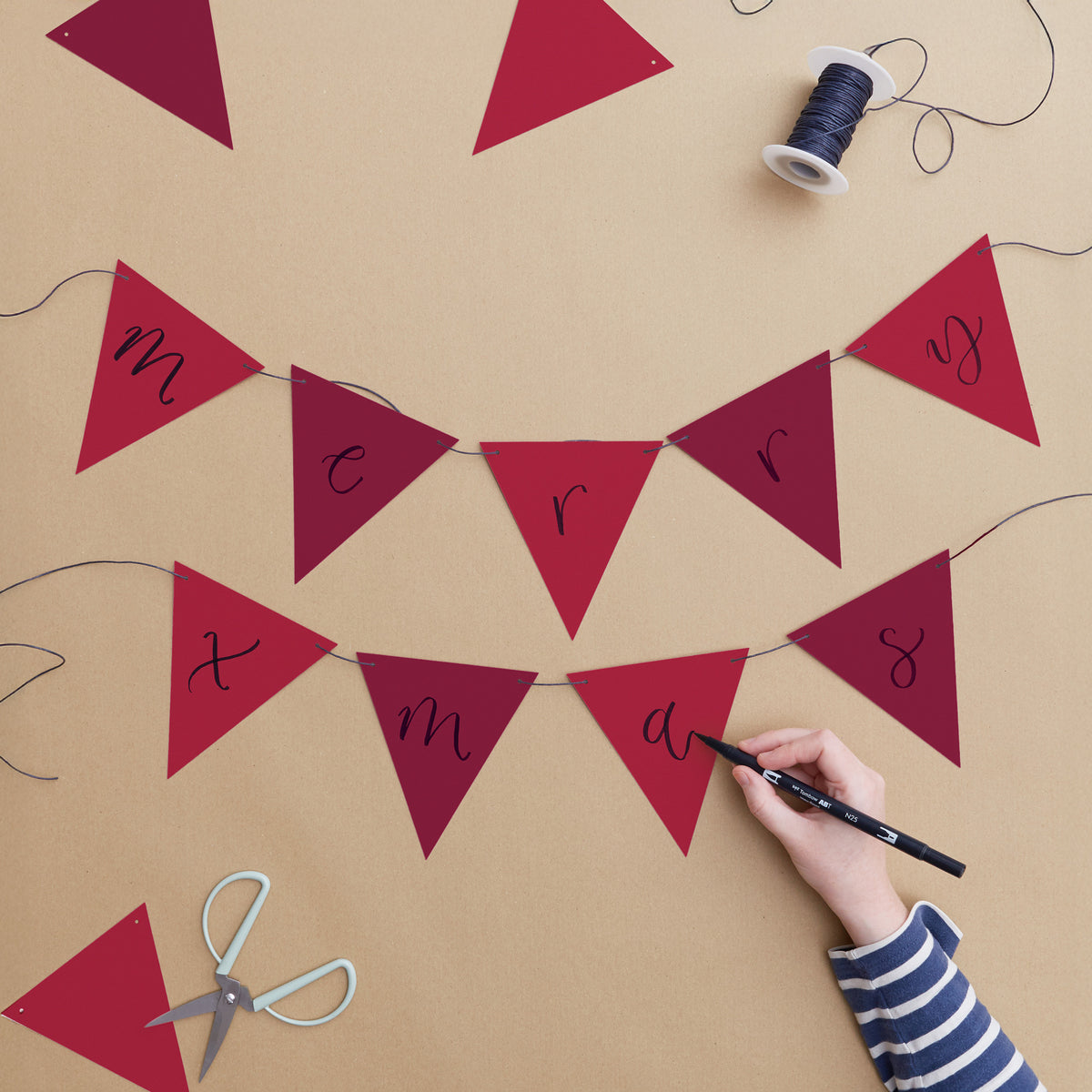 A festive Christmas bunting banner made of red triangular pennants spelling out 'Merry Christmas' on a tan background, with a hand writing on one pennant, plus scissors, thread, and extra pennants as props.