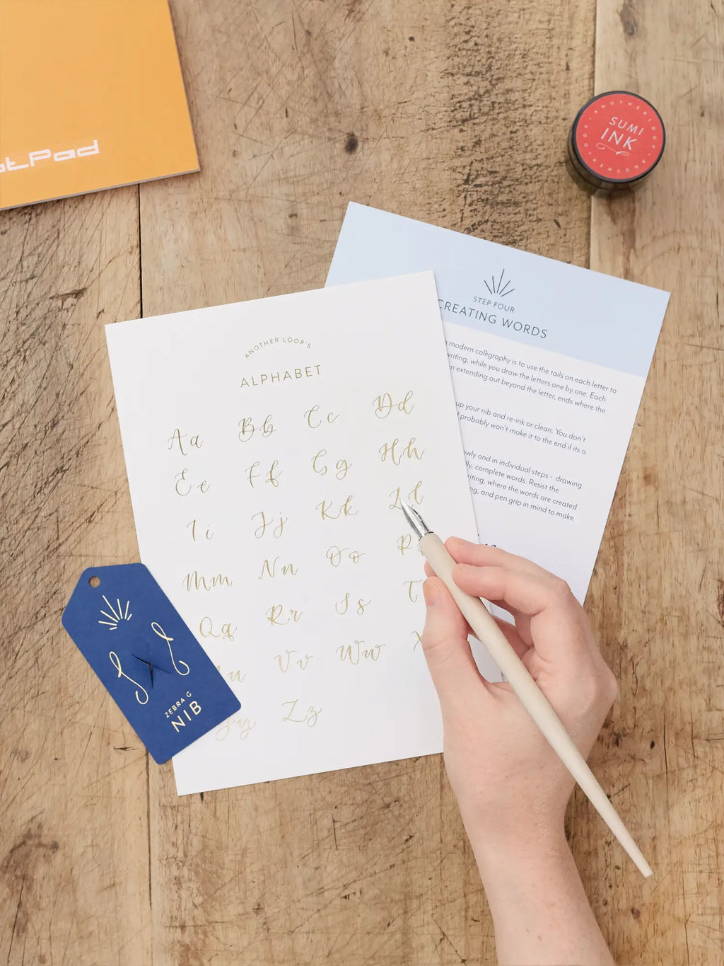 A hand holding a wooden calligraphy pen over printed alphabet practice sheets on a rustic wood table