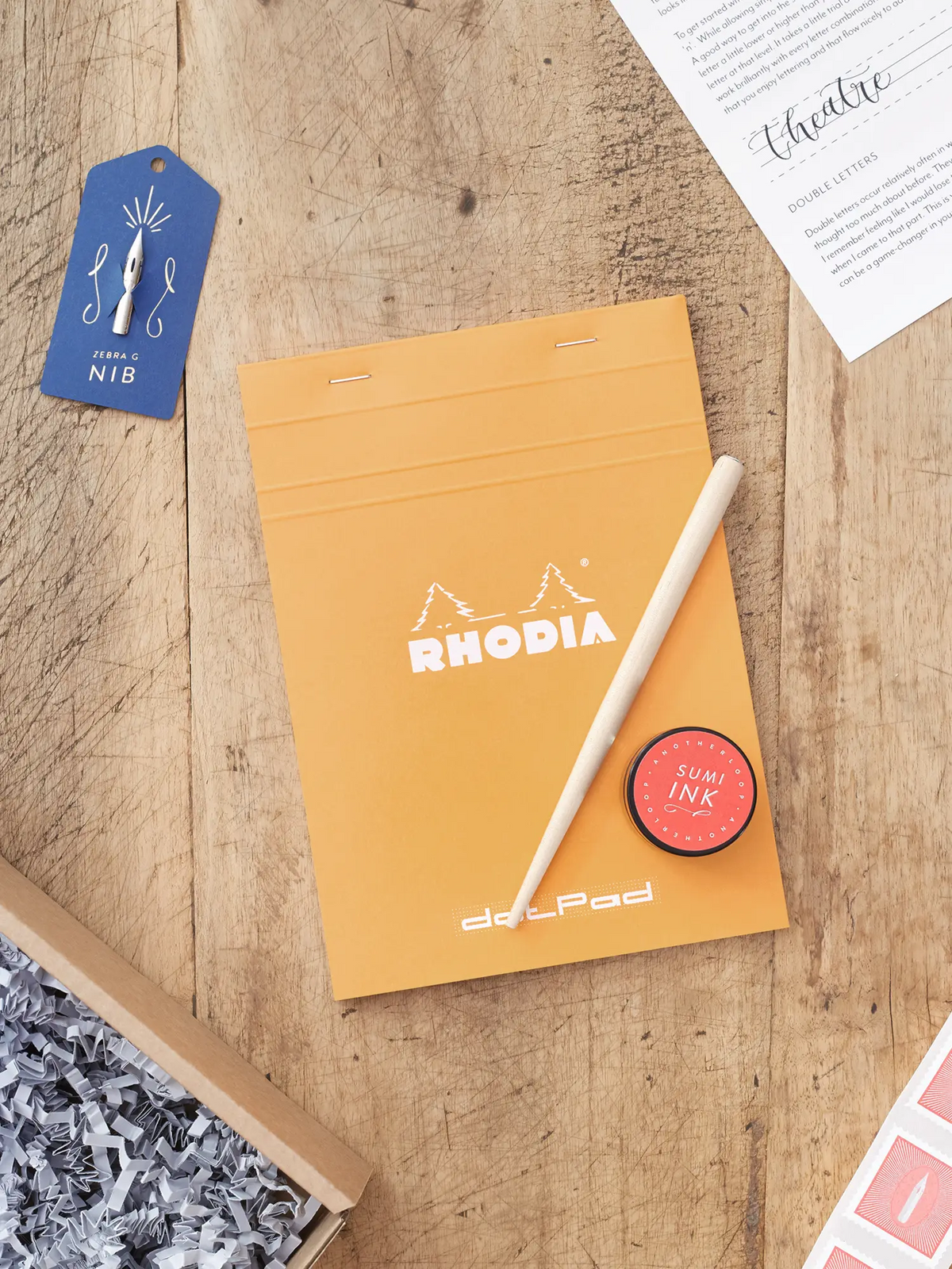 The contents of a modern calligraphy kit laid out on a rustic wooden table. At the centre is an orange Rhodia pad, accompanied by calligraphy nib in a blue tag, nib holder, black sumi ink pot and instruction sheet.