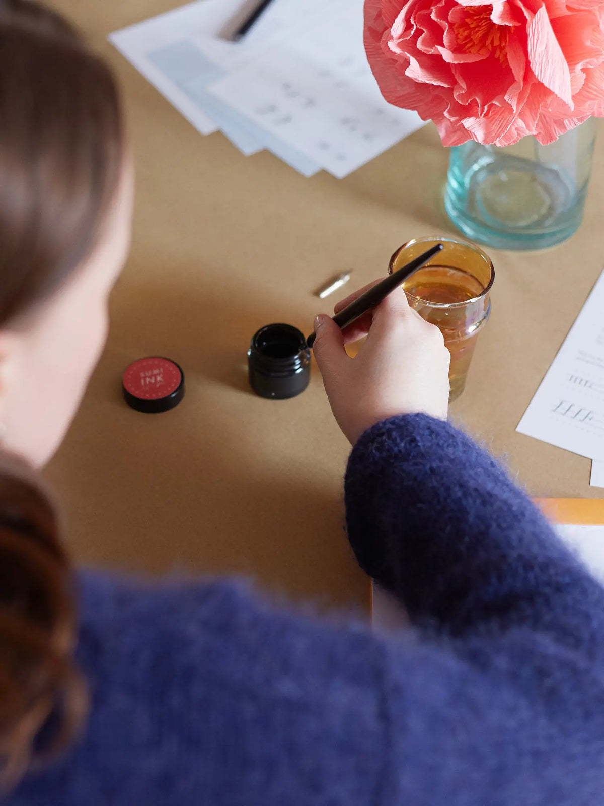 a person viewed from behind at a table in newcastle, holding a calligraphy nib over a small jar of black sumi ink