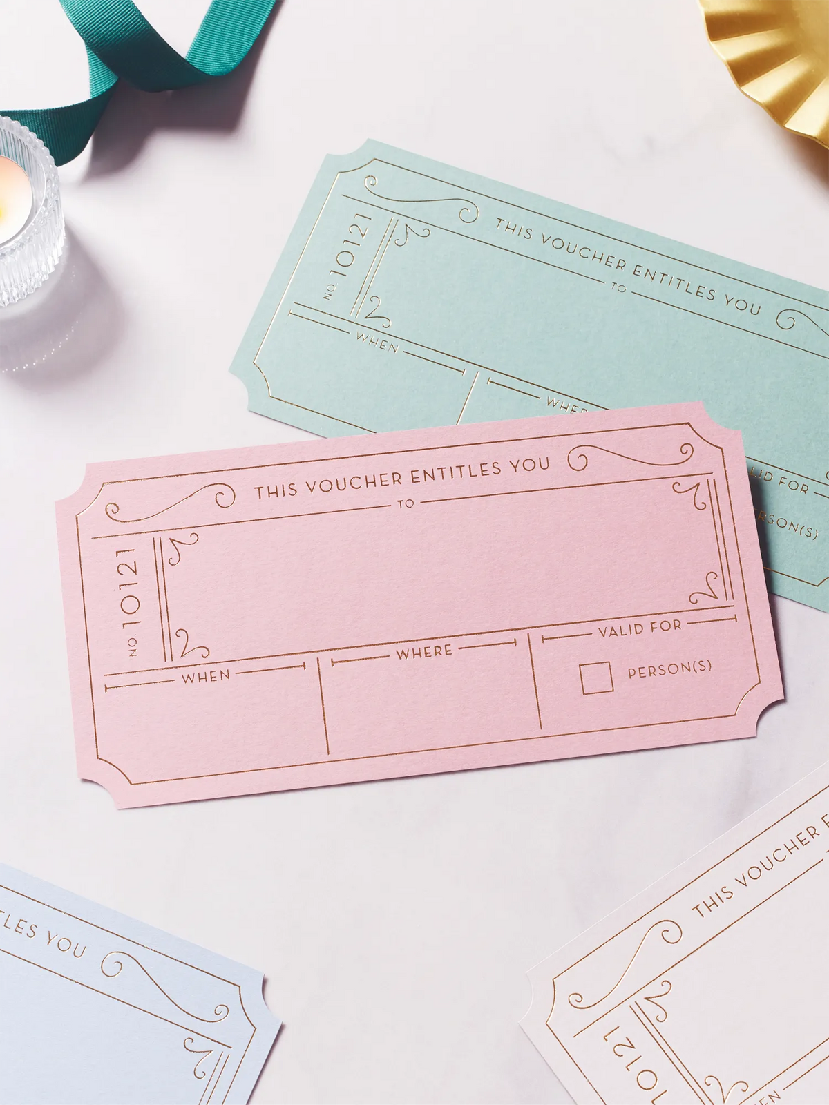 A styled flat-lay of hot foiled golden tickets in dusty pink, avocado green, chalk  blue, and plume grey, arranged on a white marble surface with ribbon and candle props.