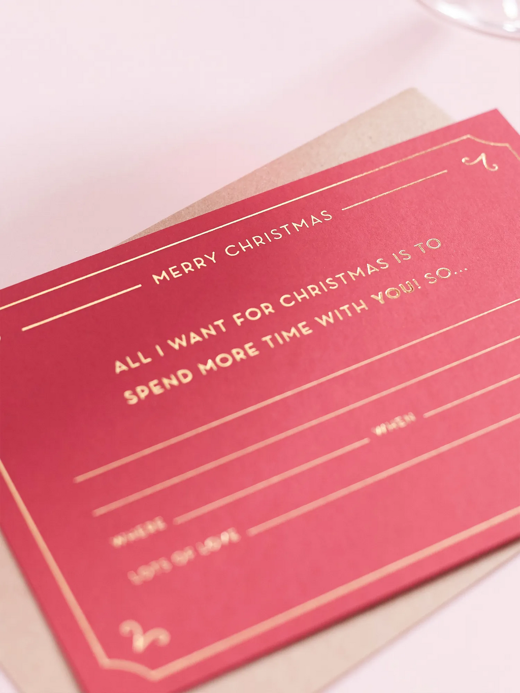 A red Christmas Gift Of Time Voucher with gold foil lettering, placed on top of a brown kraft envelope. On a pink background.