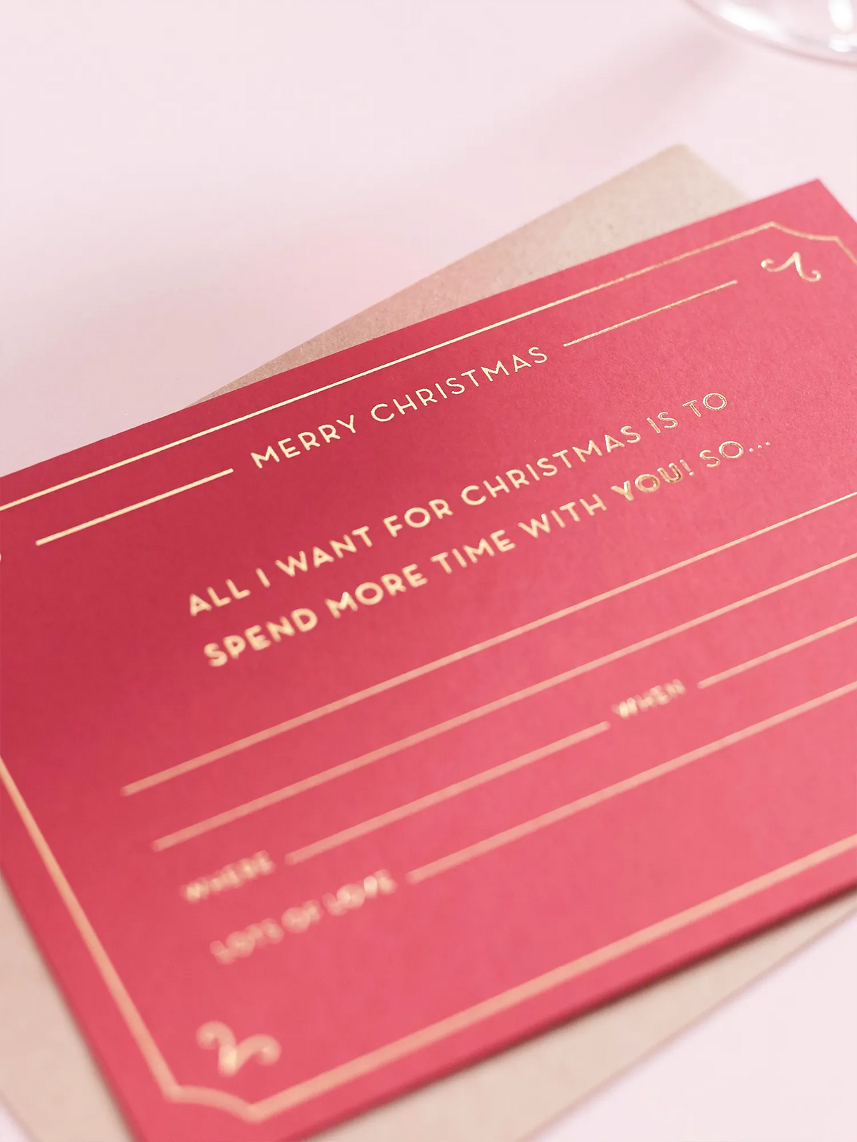 A red Christmas Gift Of Time Voucher with gold foil lettering, placed on top of a brown kraft envelope. On a pink background.