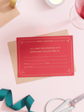 A red Christmas Gift Of Time Voucher with gold foil lettering, placed on top of a brown kraft envelope. A pair of scissors, and teal ribbon in the background on a pink surface.