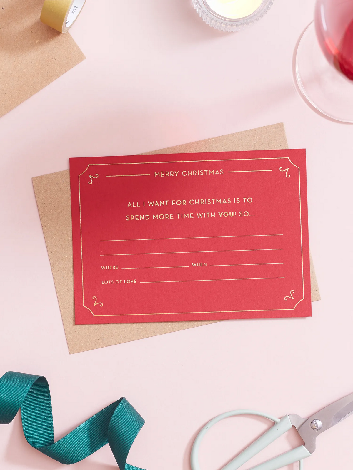 A red Christmas Gift Of Time Voucher with gold foil lettering, placed on top of a brown kraft envelope. A pair of scissors, and teal ribbon in the background on a pink surface.