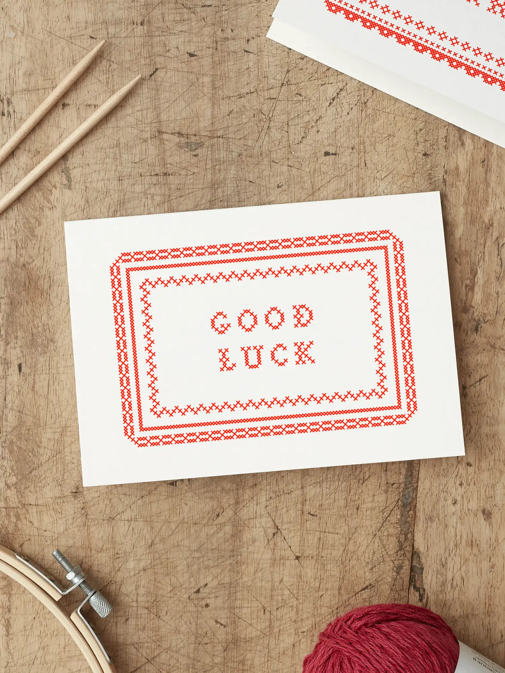 Red and White Screen printed good luck card with ‘Good Luck’ with a cross stitch folk art style illustration, placed on a wooden surface.