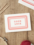 Red and White Screen printed good luck card with ‘Good Luck’ with a cross stitch folk art style illustration, placed on a wooden surface.