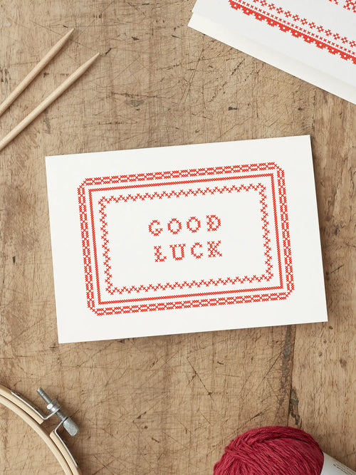 Red and White Screen printed good luck card with ‘Good Luck’ with a cross stitch folk art style illustration, placed on a wooden surface.