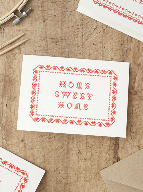 Screen printed New Home card with ‘Home Sweet Home’ with a cross stitch folk art style illustration, placed on a wooden surface.