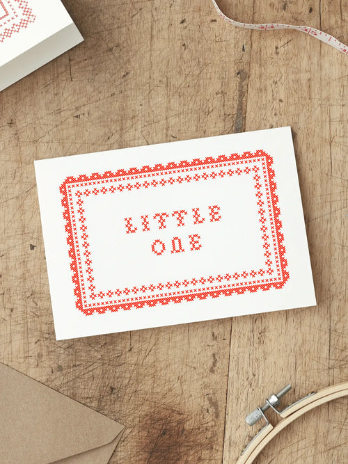 Red and White Screen printed New Baby card with ‘Little One’ with a cross stitch folk art style illustration, placed on a wooden surface.