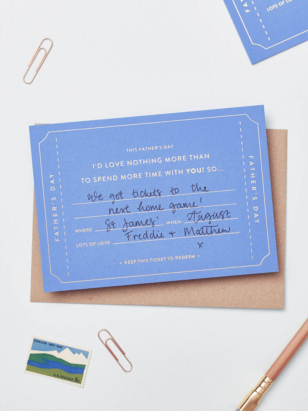 A blue Father's Day Gift Of Time Voucher with gold foil lettering filled in with black hand written text, placed on top of a brown kraft envelope. A copper paperclip and a small postage stamp are visible in the background on a white surface.