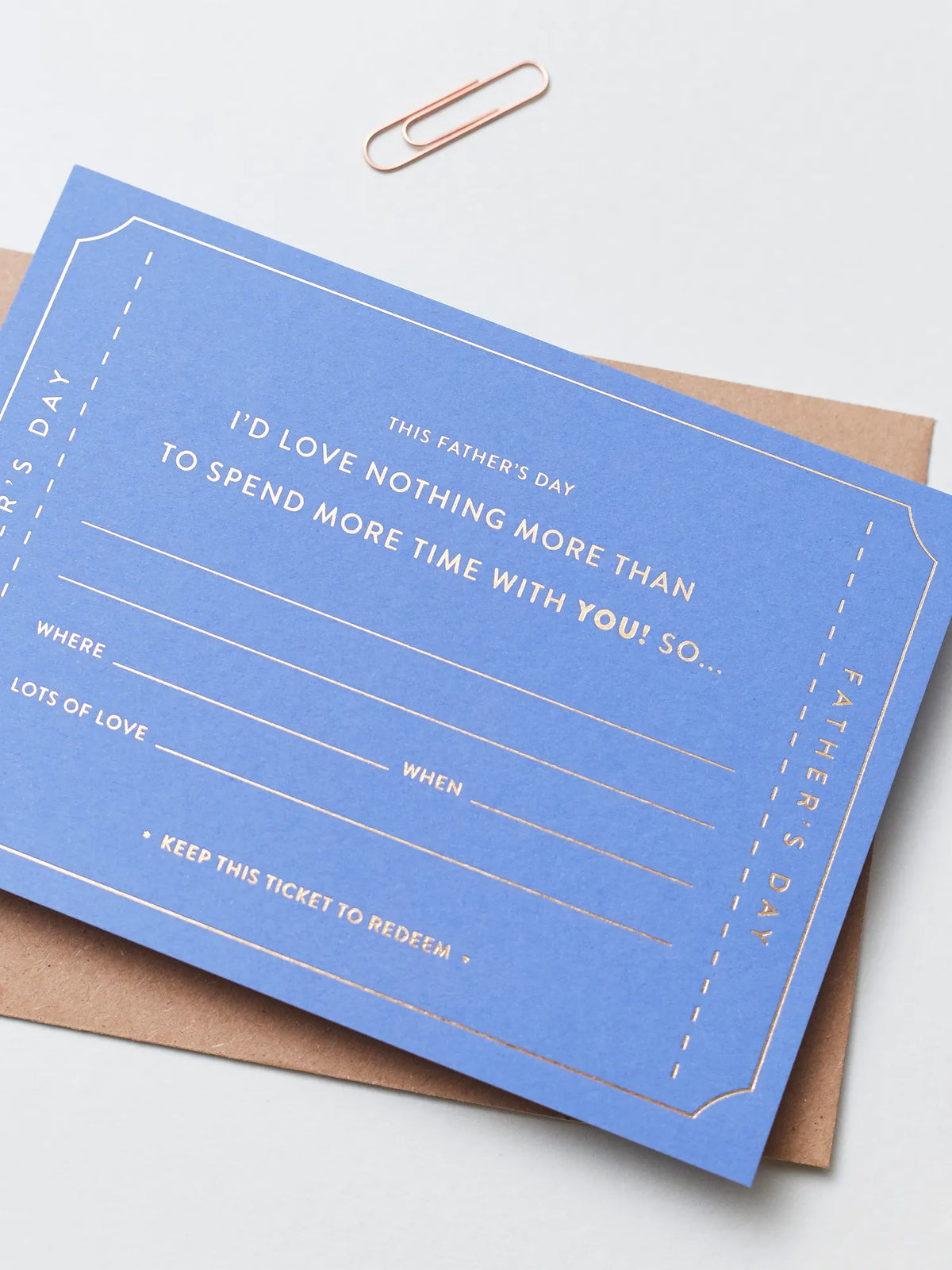 A blue Father's Day Gift Of Time Voucher with gold foil lettering, placed on top of a brown kraft envelope. A copper paperclip are visible in the background on a white surface.