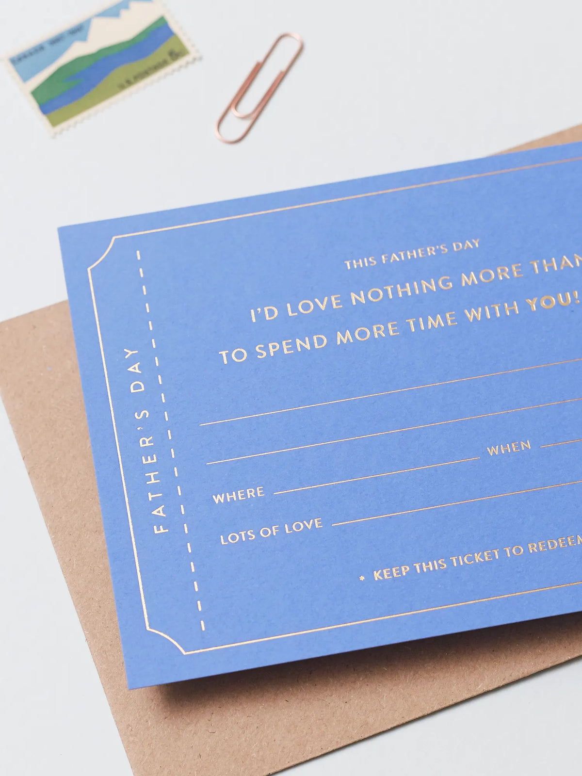A blue Father's Day Gift Of Time Voucher with gold foil lettering, placed on top of a brown kraft envelope. A copper paperclip and a small postage stamp are visible in the background on a white surface.