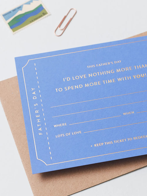A blue Father's Day Gift Of Time Voucher with gold foil lettering, placed on top of a brown kraft envelope. A copper paperclip and a small postage stamp are visible in the background on a white surface.