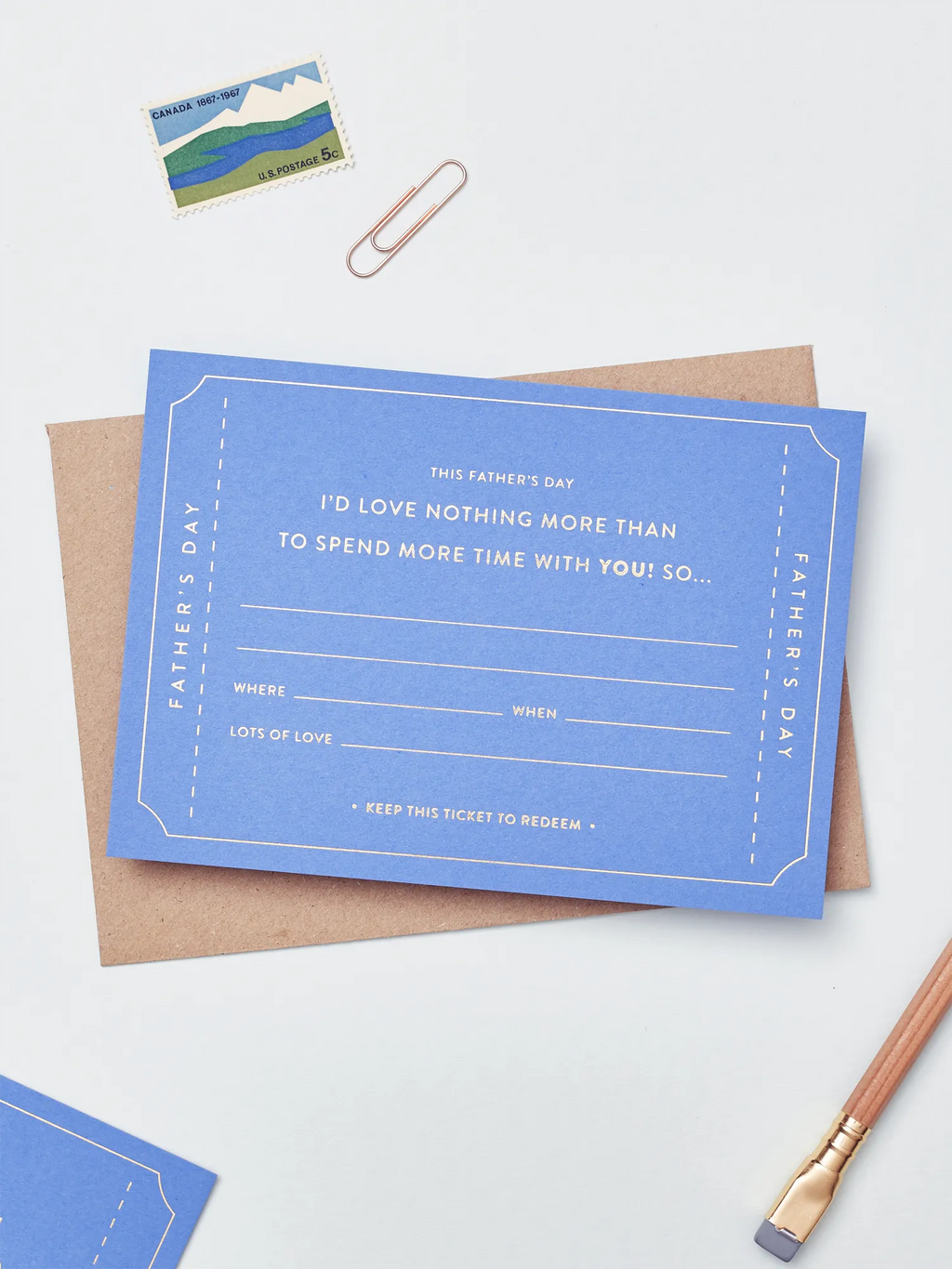 A blue Father's Day Gift Of Time Voucher with gold foil lettering, placed on top of a brown kraft envelope. A copper paperclip and a small postage stamp are visible in the background on a white surface.