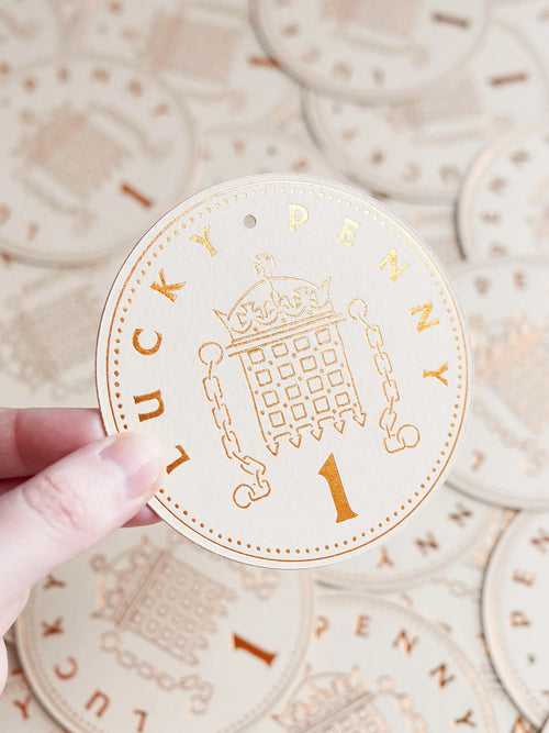 A hand holds a round, brown lucky penny token and the word 'LUCKY PENNY' around the edge, with other similar tokens scattered in the background.
