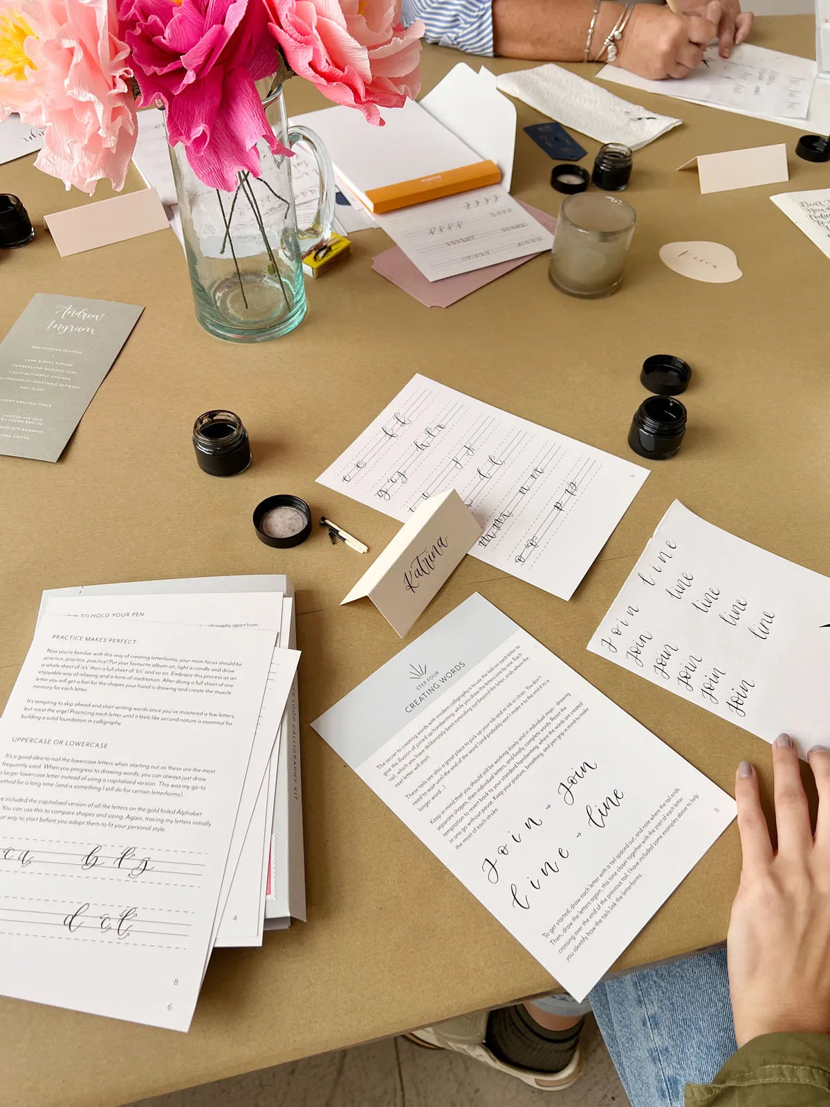 a busy calligraphy workshop table in newcastle with printed practice sheets, ink bottles, a glass vase of pink flowers and hands working on writing