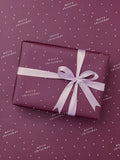 A square gift box wrapped in Burgundy paper with a lavender ribbon and bow, placed on a matching burgundy background with small white stars and ‘Merry Christmas' text.