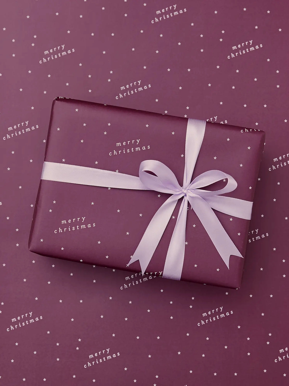 A square gift box wrapped in Burgundy paper with a lavender ribbon and bow, placed on a matching burgundy background with small white stars and ‘Merry Christmas' text.