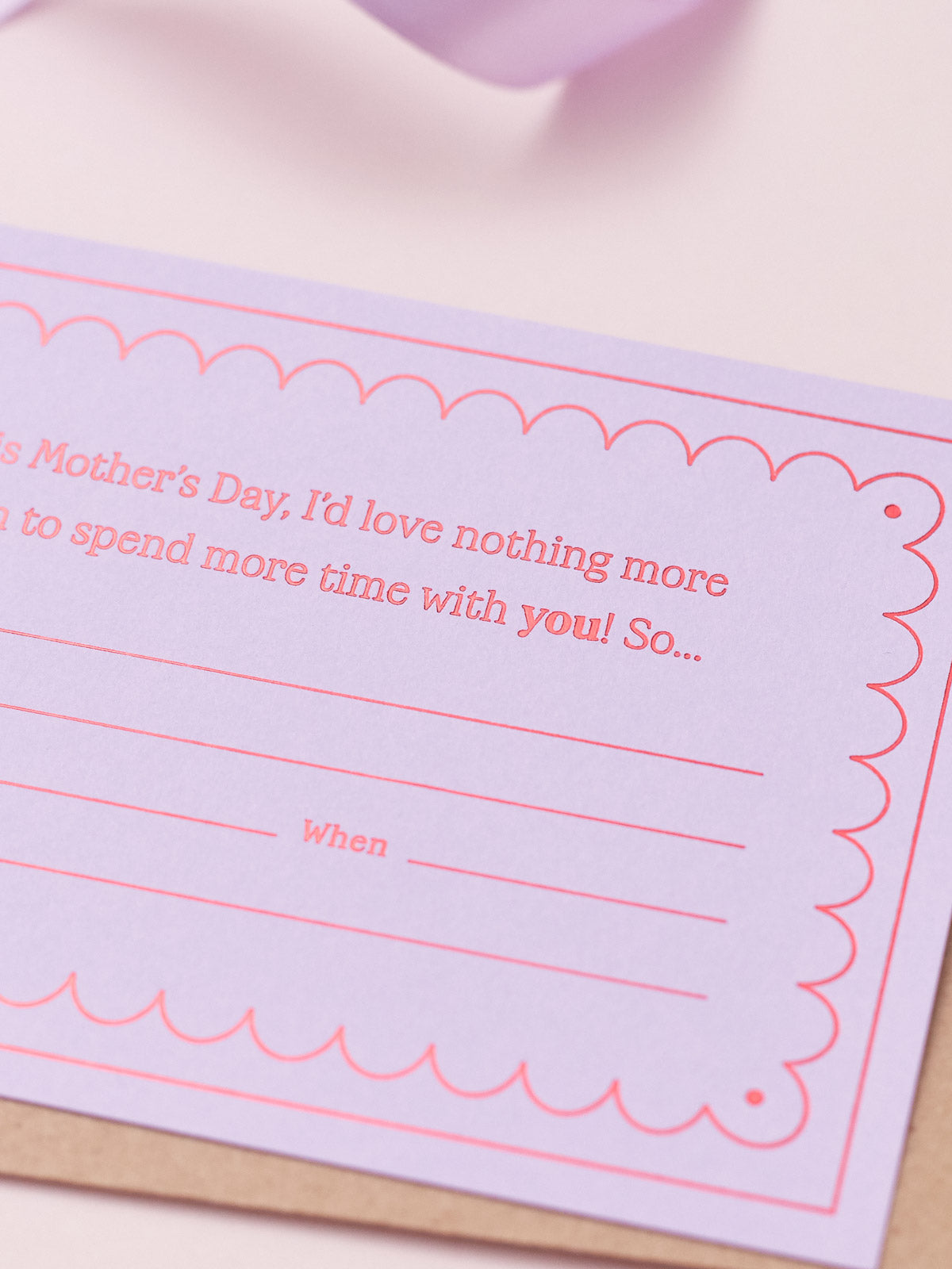 A Lavender Mother's Day Gift Of Time Voucher with red foil lettering, placed on top of a brown kraft envelope. A lavender ribbon is visible in the background on a pink surface.