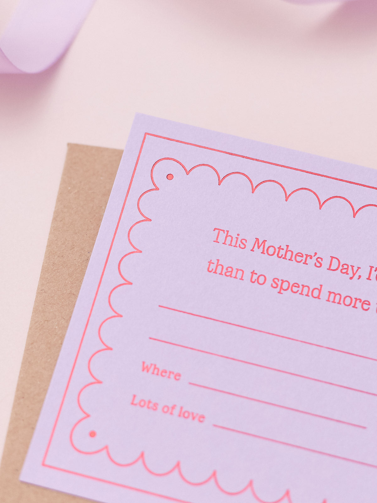 A Lavender Mother's Day Gift Of Time Voucher with red foil lettering, placed on top of a brown kraft envelope. A lavender ribbon is visible in the background on a pink surface.