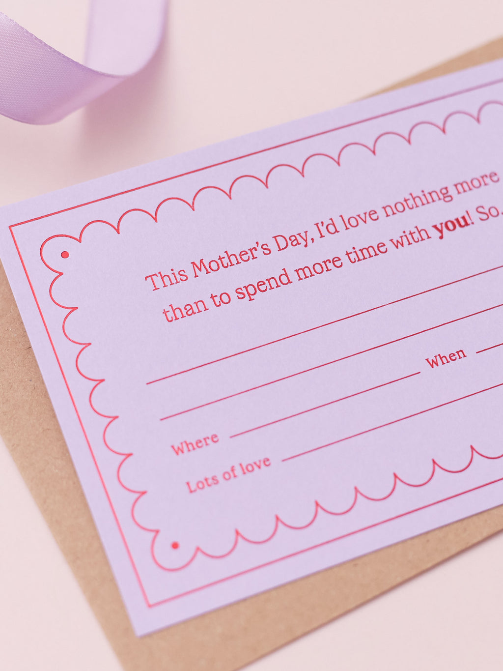 A Lavender Mother's Day Gift Of Time Voucher with red foil lettering, placed on top of a brown kraft envelope. A lavender ribbon is visible in the background on a white surface.