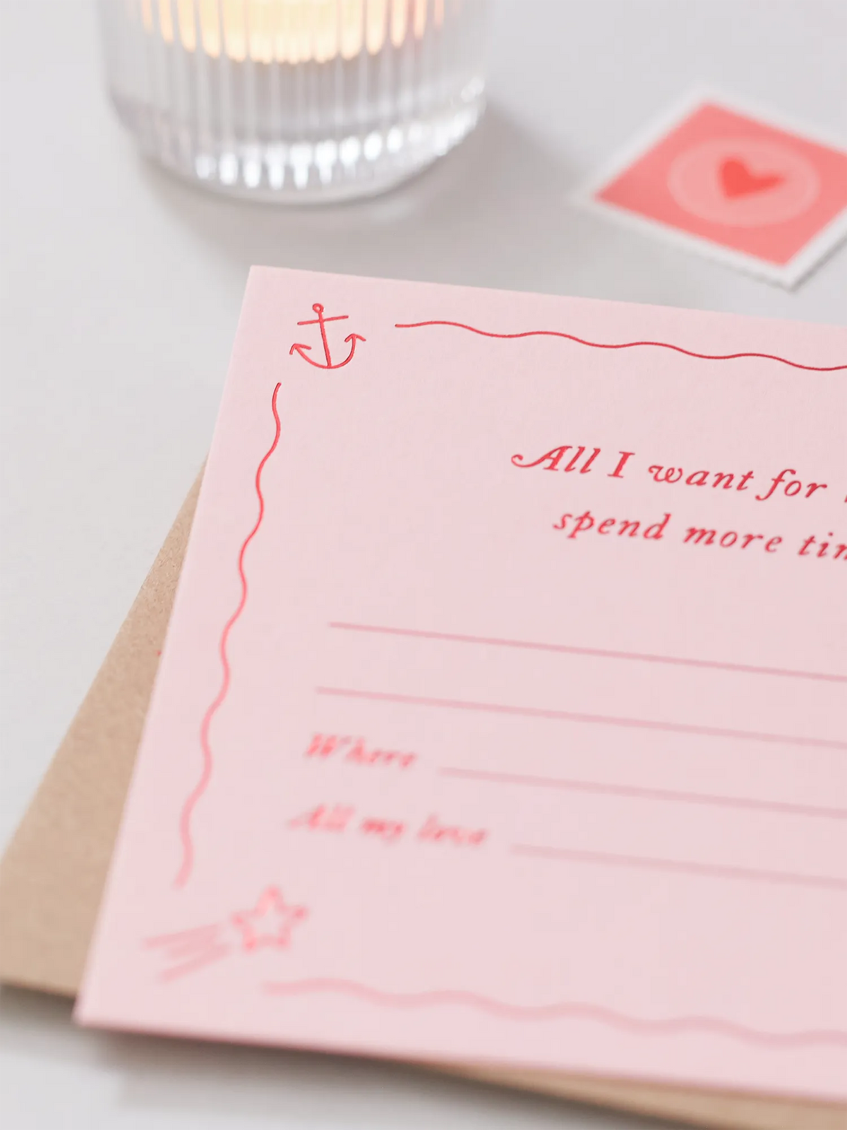 A pink Valentines's Day Gift Of Time Voucher with red foil lettering, placed on top of a brown kraft envelope. A heart stamp is visible in the background on a grey surface.
