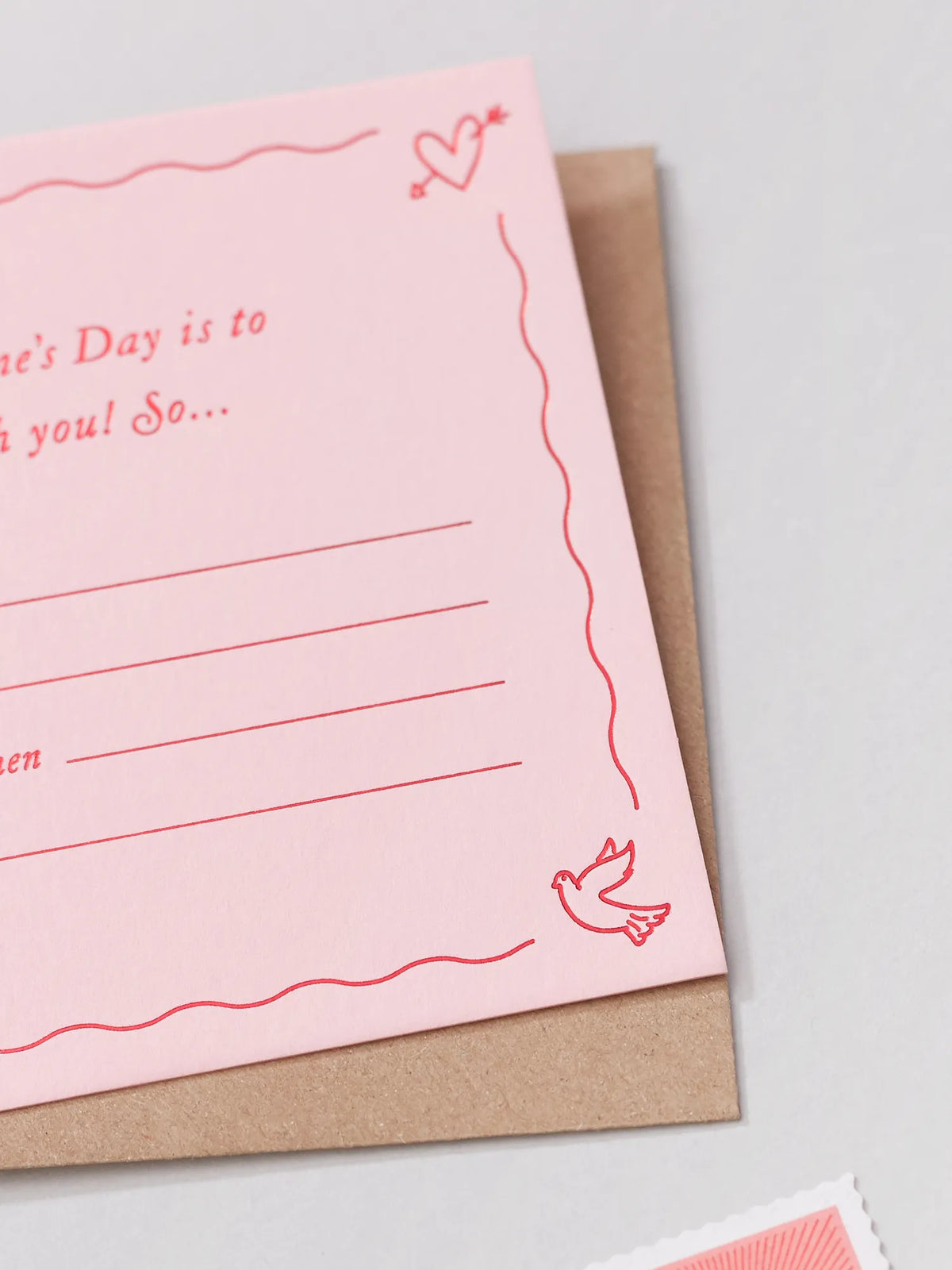 A pink valentines Day Gift Of Time Voucher with red foil lettering, placed on top of a brown kraft envelope. A close up of an illustrated dove in the bottom corner