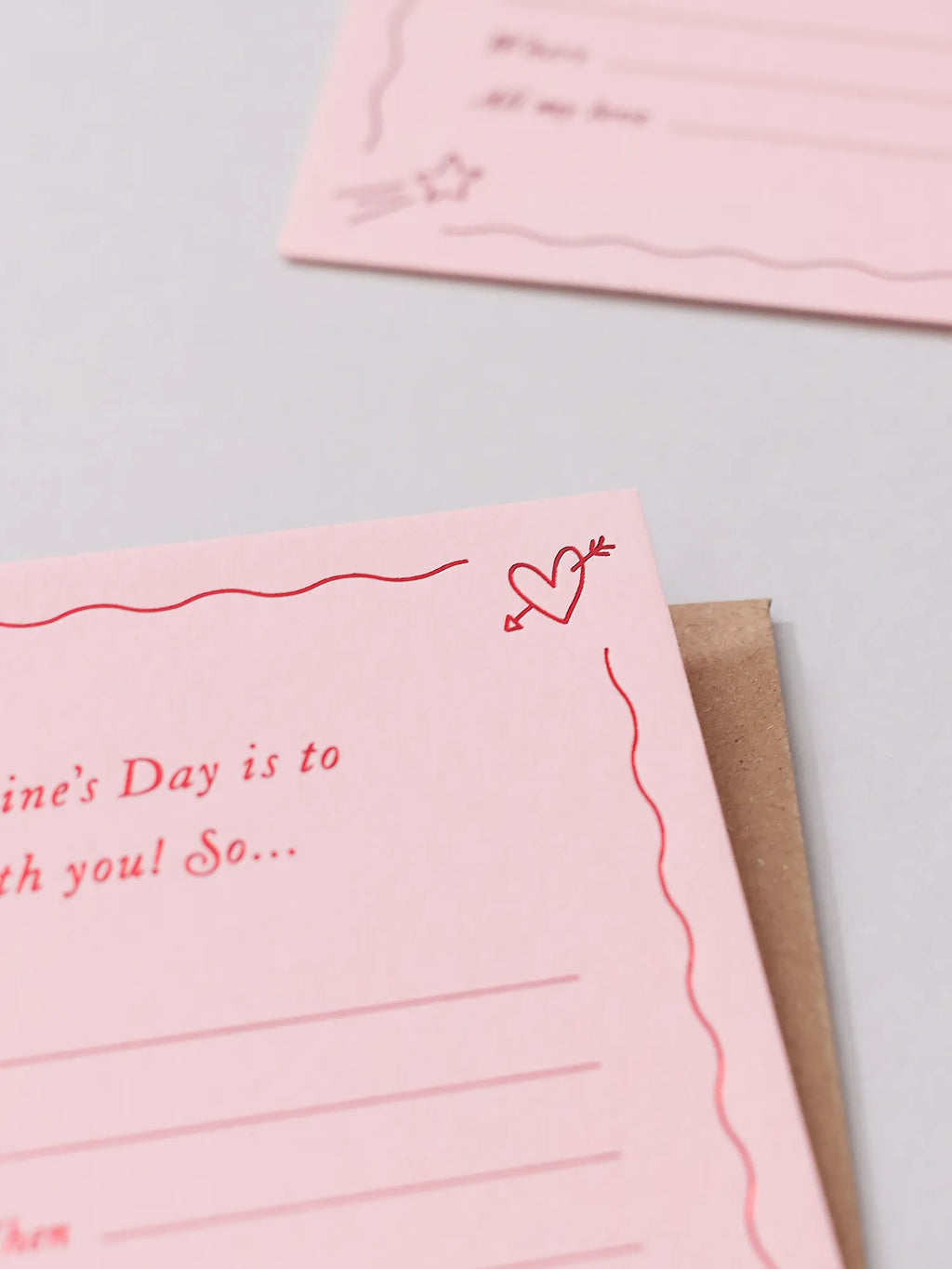 A pink Valentine's Day Gift Of Time Voucher with red foil lettering, placed on top of a brown kraft envelope. close up of a love heart with arrow through