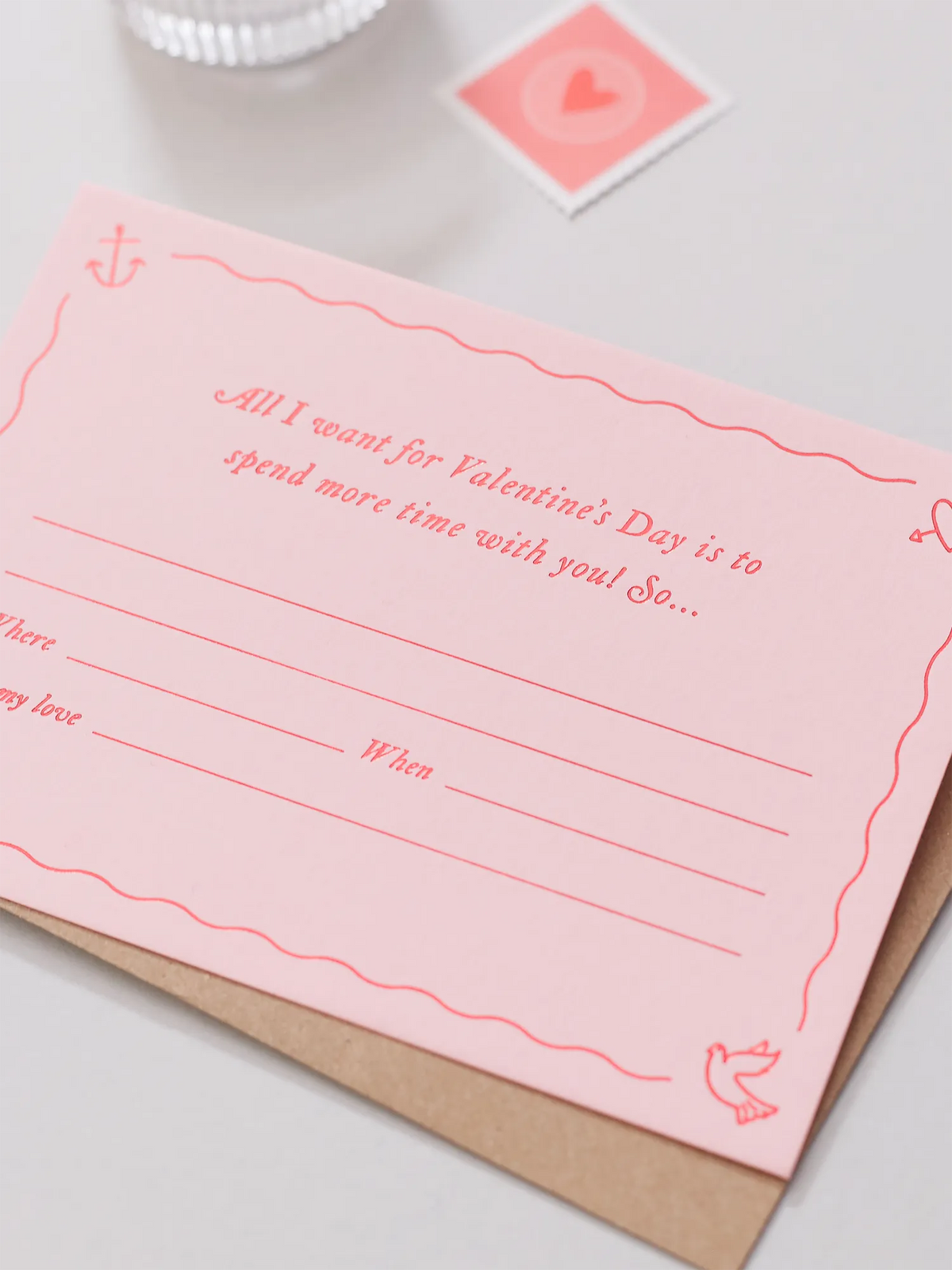 A pink Valentines Day Gift Of Time Voucher with red foil lettering, placed on top of a brown kraft envelope. A love heart stamp is visible in the background on a grey surface.