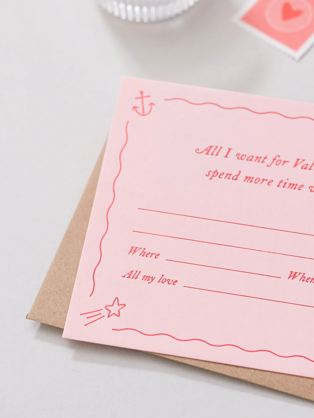 A pink Valentines Day Gift Of Time Voucher with red foil lettering, placed on top of a brown kraft envelope. A love heart stamp is visible in the background on a grey surface.