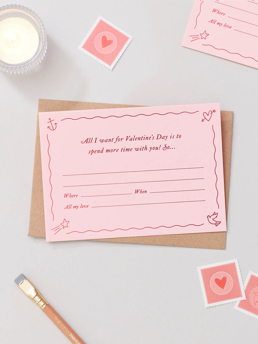 A pink Valentines Day Gift Of Time Voucher with red foil lettering, placed on top of a brown kraft envelope. A love heart stamp scattered around in the background on a grey surface.