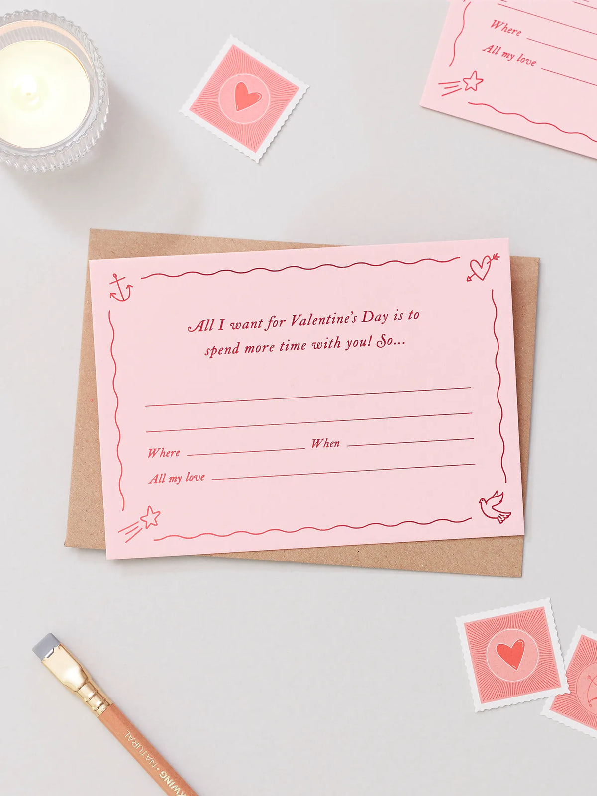 A pink Valentines Day Gift Of Time Voucher with red foil lettering, placed on top of a brown kraft envelope. A love heart stamp scattered around in the background on a grey surface.